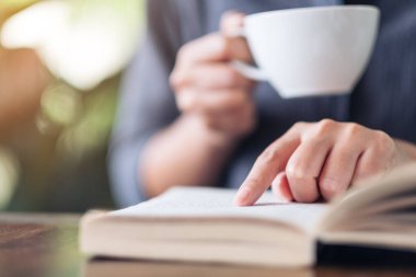 Ahşap masada kahve içerken kitap okuyup işaret eden bir kadının yakın plan resmi.
