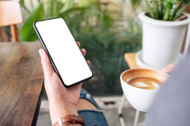 Kafede kahve içerken elinde siyah cep telefonu ve boş masaüstü ekran kullanan bir kadının görüntüsü.