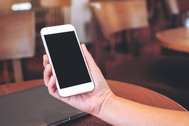 Masanın üzerinde dizüstü bilgisayar ve boş masaüstü ekranı olan beyaz cep telefonu tutan ellerin görüntüsü