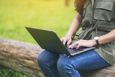 Parktaki bir kütüğün üzerinde otururken dizüstü bilgisayarda yazan ve kullanan bir kadın.