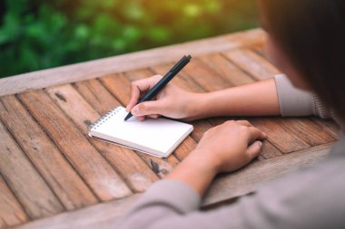 Kapalı alanda ahşap masaya boş bir deftere yazı yazan bir kadının yakın plan resmi.