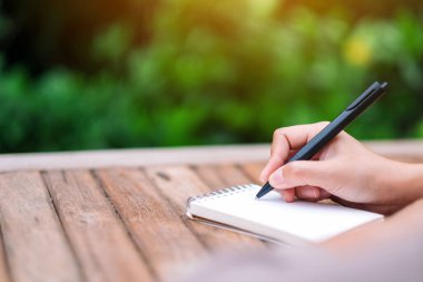 Kapalı alanda ahşap masaya boş bir deftere yazı yazan bir kadının yakın plan resmi.