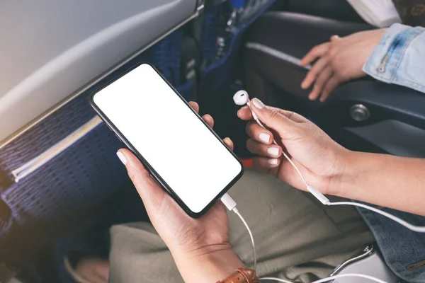 Mockup image of woman's hand holding a black smart phone with blank desktop screen and earphone in cabin