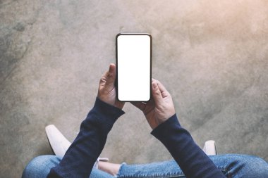 Üst görünümde, yerde otururken elinde beyaz ekranlı siyah bir cep telefonu tutan bir kadının görüntüsü var.