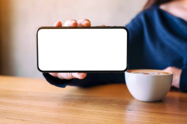 Kahve içerken elinde siyah bir cep telefonu tutan ve gösteren beyaz ekranlı bir kadının görüntüsü.