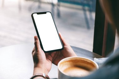 Elinde siyah cep telefonu olan, boş ekranlı, ahşap sehpada kahve fincanı olan bir kadının görüntüsü. 