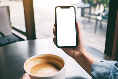 Masada kahve içerken elinde boş ekranla siyah cep telefonu tutan bir kadının görüntüsü. 