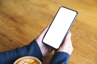 Beyaz cep telefonlu, boş ekranlı, ahşap masada kahve fincanı olan bir kadının görüntüsü. 