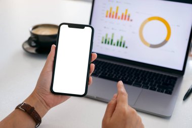 Ofiste dizüstü bilgisayar kullanırken siyah bir cep telefonunu boş ekranla tutan ellerin görüntüsü.