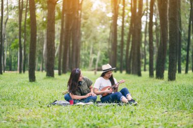 İki kadın ormanda oturup rahatlarken ukulele oynuyor.