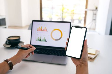 Ofiste dizüstü bilgisayar kullanırken siyah bir cep telefonunu boş ekran ile tutan bir elin görüntüsü