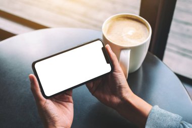 Siyah cep telefonlu, boş ekranlı ve ahşap masada kahve fincanı olan ellerin görüntüsü. 