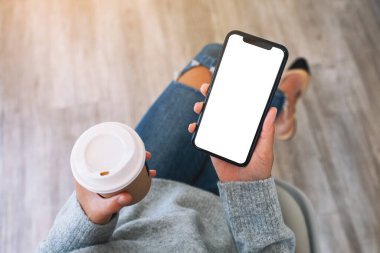 Üst görünümde, elinde siyah bir cep telefonu olan bir kadının kahve fincanı olan beyaz ekranlı görüntüsü var.