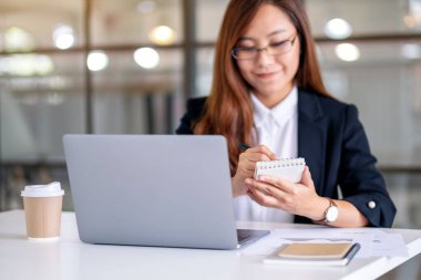 Ofiste dizüstü bilgisayarla çalışırken not defterine yazan iş kadınının yakın plan resmi