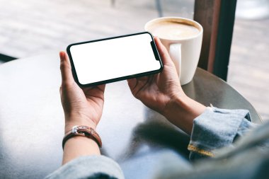 Siyah cep telefonlu, boş ekranlı ve ahşap masada kahve fincanı olan ellerin görüntüsü. 