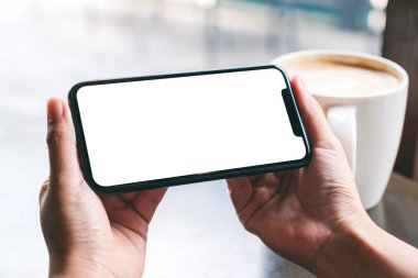 Siyah cep telefonlu, boş ekranlı ve ahşap masada kahve fincanı olan ellerin görüntüsü. 