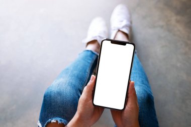 Üst görünümde, yerde otururken elinde beyaz ekranlı siyah bir cep telefonu tutan bir kadının görüntüsü var.