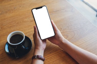 Elinde siyah cep telefonu olan, boş ekranlı, ahşap sehpada kahve fincanı olan bir kadının görüntüsü.