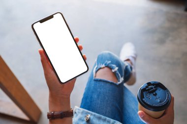 Üst görünümde, elinde siyah bir cep telefonu olan bir kadının kahve fincanı olan beyaz ekranlı görüntüsü var.