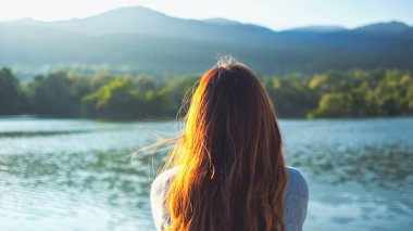 Gölün kenarında tek başına oturan bir kadın yeşil doğalı dağlara bakıyor.