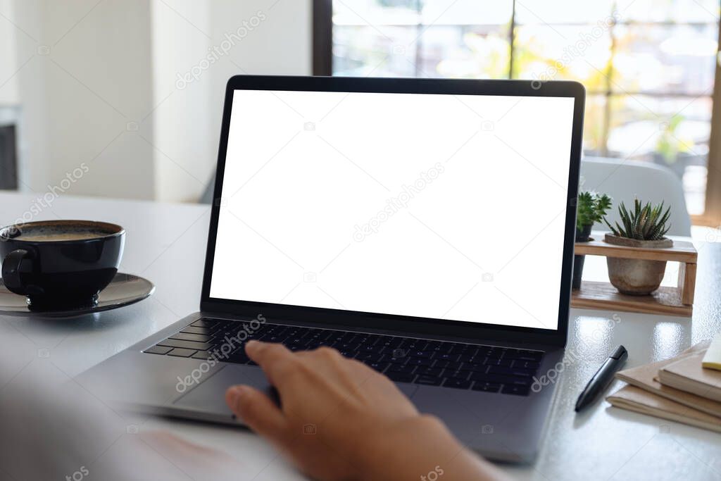 Mockup image of a hand using and touching on laptop touchpad with blank white desktop screen with coffee cup on wooden table 