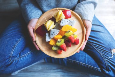 Zeminde otururken, elinde taze karışık meyve tabağı tutan ve yemek yiyen bir kadın resmi.