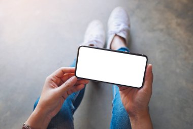 Üst görünümde, yerde otururken elinde beyaz ekranlı siyah bir cep telefonu tutan bir kadının görüntüsü var.