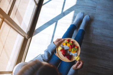 Zeminde otururken, elinde taze karışık meyve tabağı tutan ve yemek yiyen bir kadın resmi.