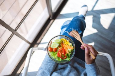 Bir sandalyede otururken elinde taze sebze salatası tutan bir kadının görüntüsü.