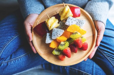 Bir kadının ahşap bir tabak dolusu taze karışık meyveyi yerde otururken görüntüsü.