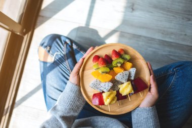 Bir kadının ahşap bir tabak dolusu taze karışık meyveyi yerde otururken görüntüsü.