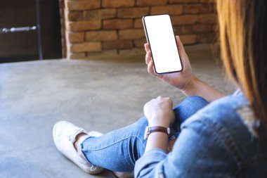 Yerde otururken elinde beyaz ekranlı siyah bir cep telefonu tutan bir kadının görüntüsü.