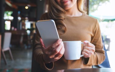 Kafede kahve içerken cep telefonu tutan ve kullanan genç bir kadının yakın plan fotoğrafı.