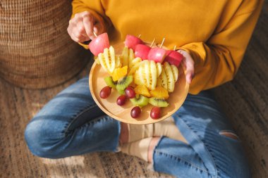 Elinde tahta bir tabak taze karışık meyve tutan bir kadının resmi.