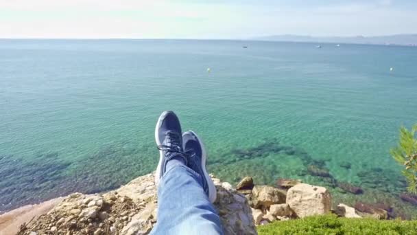 Погляд від першої особи, знятий з пішохода, що сидить на краю скелі над блакитним морем — стокове відео