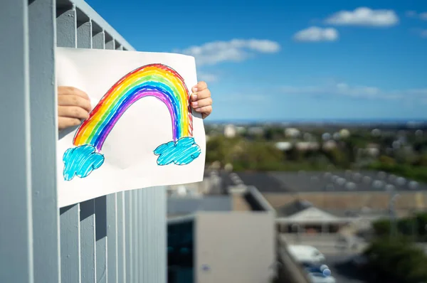 Niño sosteniendo este dibujo del arco iris en el balcón de casa. Concepto de Cuarentena del Coronavirus — Foto de Stock