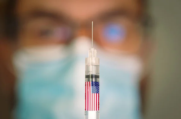 Close-up of hypodermic syringe with an USA flag and blurred doctor on the background. — Stock Photo, Image