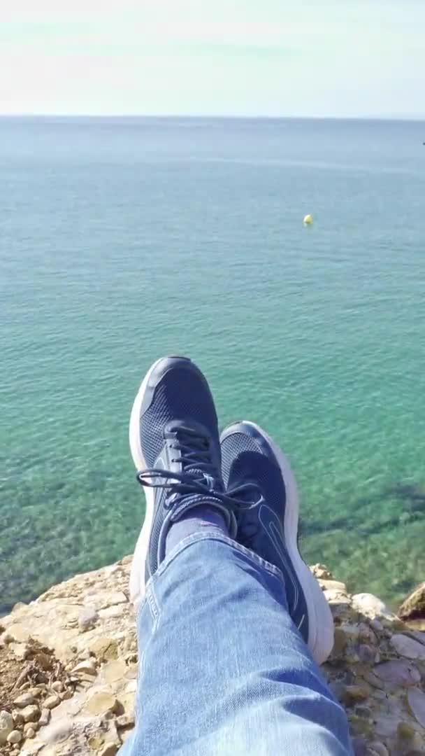 Vídeo vertical de uma perspectiva de primeira pessoa filmado sentado na borda de um penhasco sobre o mar azul — Vídeo de Stock