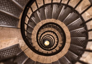 Capturing a mesmerizing view from above, a spiral staircase descends with elegant metal handrails and beige stone steps, creating a hypnotic pattern that draws the eye