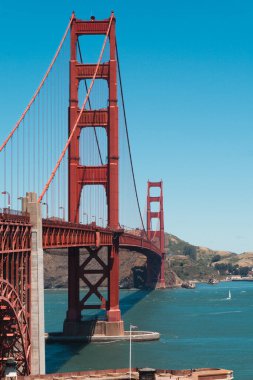 Golden Gate Köprüsü, açık güneşli gökyüzü altında mavi su üzerinde yükselen uluslararası turuncu süspansiyon kuleleri ve yeşil Marin tepeleri ile San Francisco Körfezi boyunca uzanır.