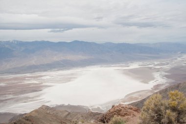 Badwater Havzaları, bulutlu bir gökyüzünün altındaki uzak dağlara doğru uzanan geniş bir çöl manzarasına hakim, aşırı kurak koşullar ve jeolojik oluşumlar sergiliyor.