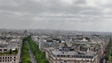 Paris şehir manzarasını yavaş yavaş kaplayan geniş hava manzarası ikonik Eyfel Kulesi, Champs Elysees Bulvarı ve bulutlu gökyüzü altındaki tarihi mimariyi gözler önüne seriyor.