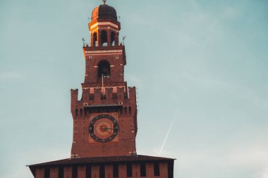 İtalya 'nın Milano kentindeki Castello Sforzesco Filarete Kulesi, İtalyan mirasını ve rönesans tasarımını temsil eden açık mavi gökyüzüne karşı tarihi tuğla mimarisini ve büyük saati gösteriyor.