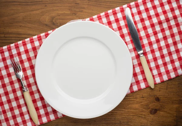 Empty plate with fork and knife on tablecloth over wooden — Stock Photo, Image