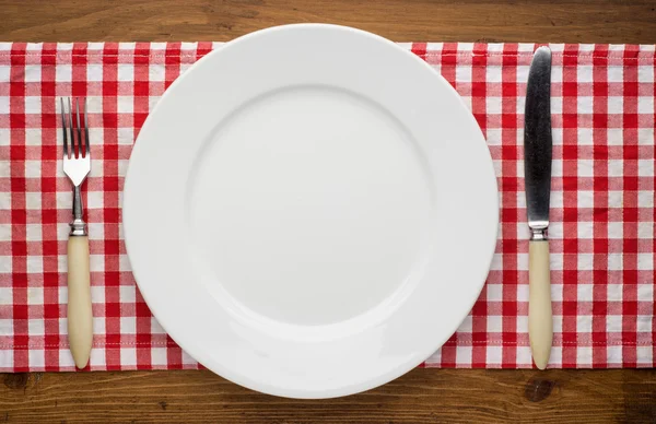 Empty plate with fork and knife on tablecloth over wooden — Stock Photo, Image