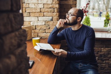 bir kafede oturan ve bir kitap okuma gözlüklü genç bir adam
