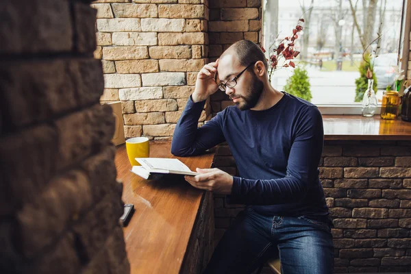 Un jeune homme avec des lunettes assis dans un café et lisant un livre — Photo