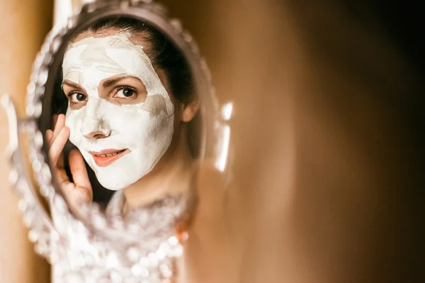 Frau trägt Maske aus Ton auf Gesicht auf und schaut in Spiegel. — Stockfoto