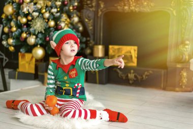 Evde Noel ağacı süsleyen çocuk. Elf kostümlü ve şapkalı küçük bir çocuk. Çocuklu aile kış tatilini kutlar. Çocuklar Noel için oturma odasını ve şömineyi dekore ederler..