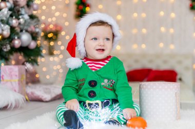 Noel hediyeleri ve oyuncaklarıyla elf gibi giyinmiş bir çocuk. Zarif bir Noel ağacının yanında hediye kutusu ve şekeri olan küçük bir çocuk. Aile kış tatilini kutluyor. Ev dekorasyonu.